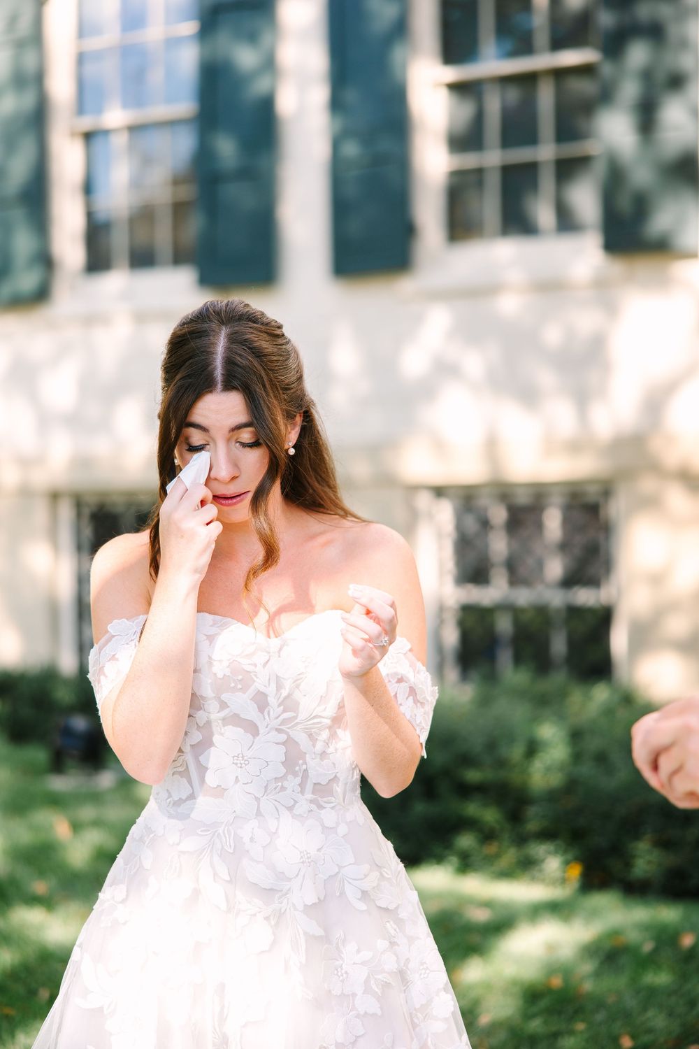 An emotional bride in a white lace off-shoulder wedding dress wipes away tears during an outdoor photo session.
