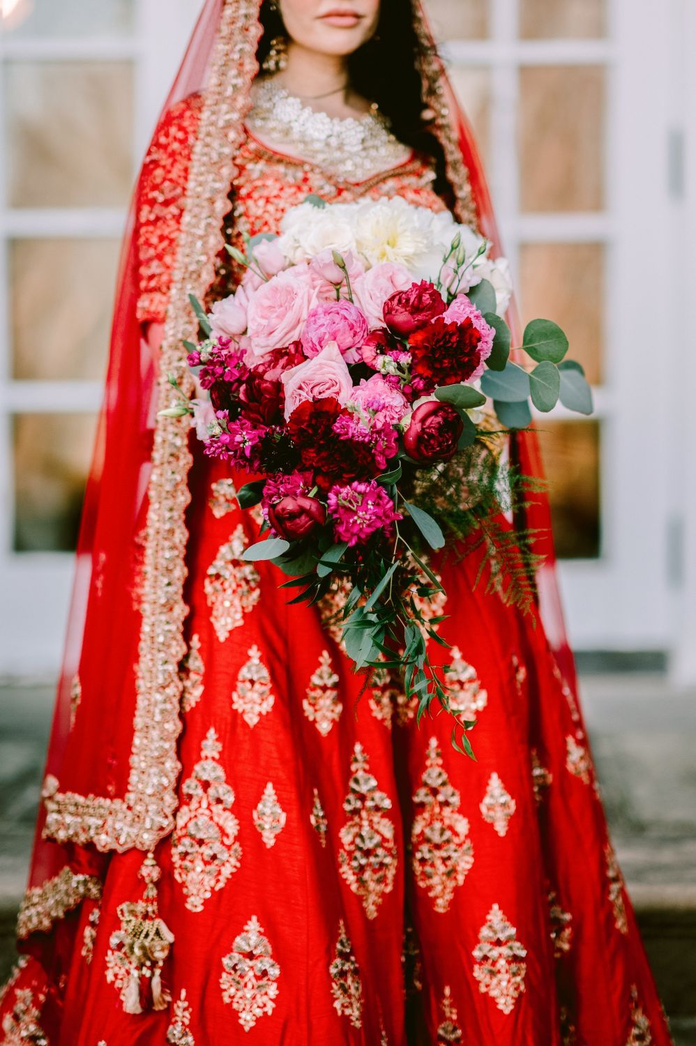 Traditional South Asian bride in red embroidered lehenga holds vibrant pink and burgundy floral bouquet with eucalyptus.
