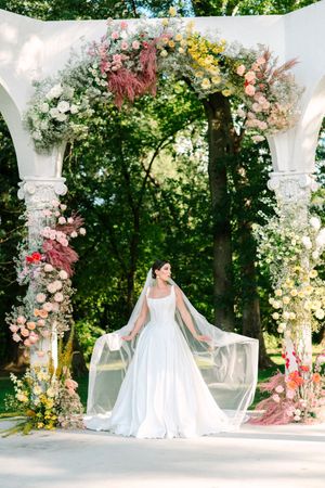 A floral wedding arch decorated with pink, yellow and white flowers frames a scenic outdoor ceremony setting.
