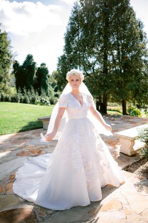 A bride in a flowing white ball gown twirls gracefully on a stone patio surrounded by lush greenery.