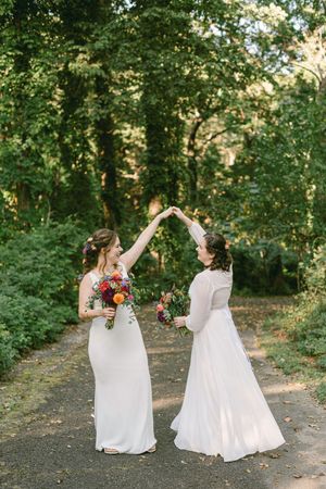 Two people in white wedding dresses dance together on a wooded path while holding colorful bouquets.
