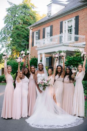 Wedding party in blush pink bridesmaid dresses poses together outside a brick building on a sunny day.