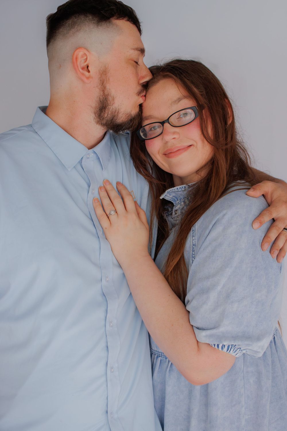 A man in a light blue shirt is kissing a woman with glasses on the cheek.