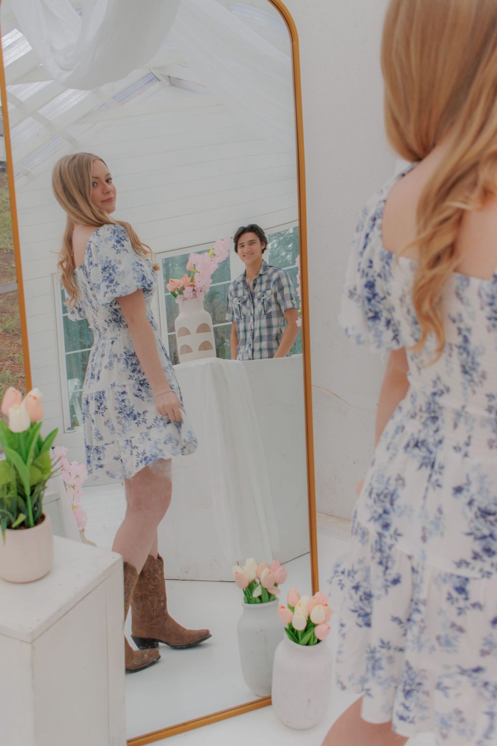 A young woman wearing a floral print dress and cowboy boots is looking at her reflection in a mirror.