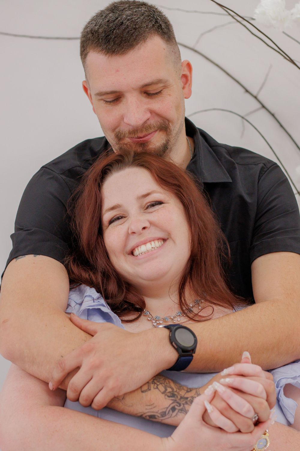 A smiling couple embracing warmly, man in black shirt hugging woman with red hair and tattoos, holding hands.