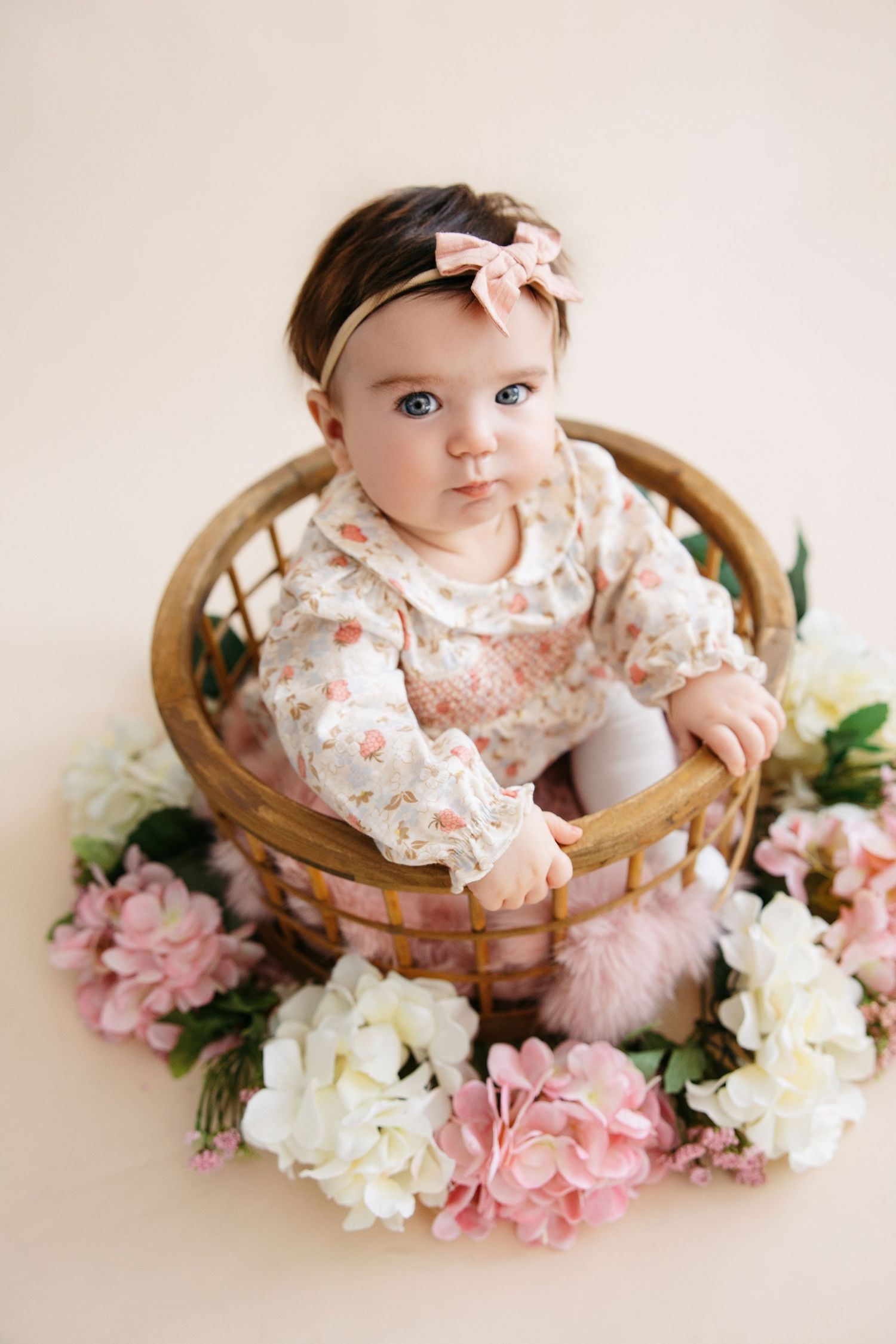 A baby sits in a wooden basket surrounded by pink and white flowers for a spring portrait session.