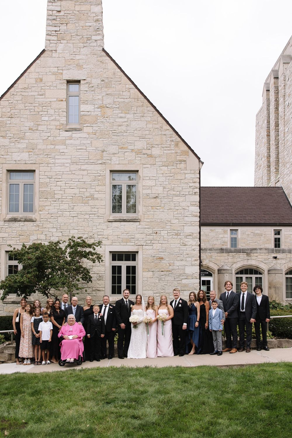 A large group of wedding guests pose for a panoramic photo outside a stone church building.