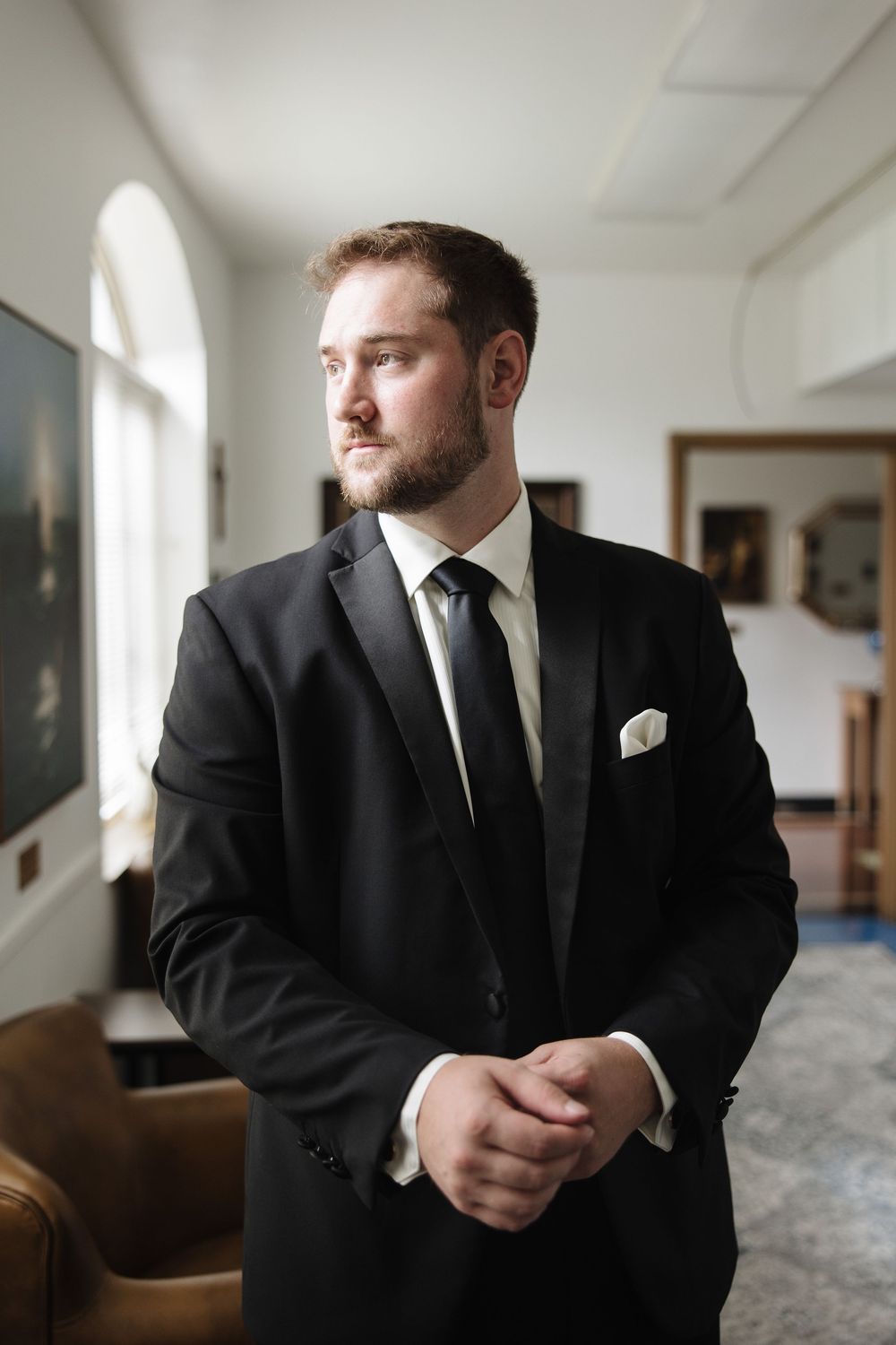 A person in a formal black suit and tie stands thoughtfully in an elegant room with arched windows.