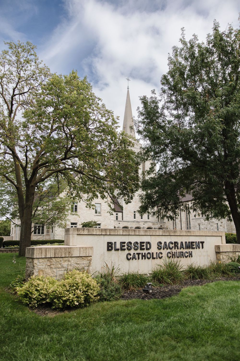 White church steeple and sign for Blessed Sacrament Catholic Church visible through trees on a grassy lawn.