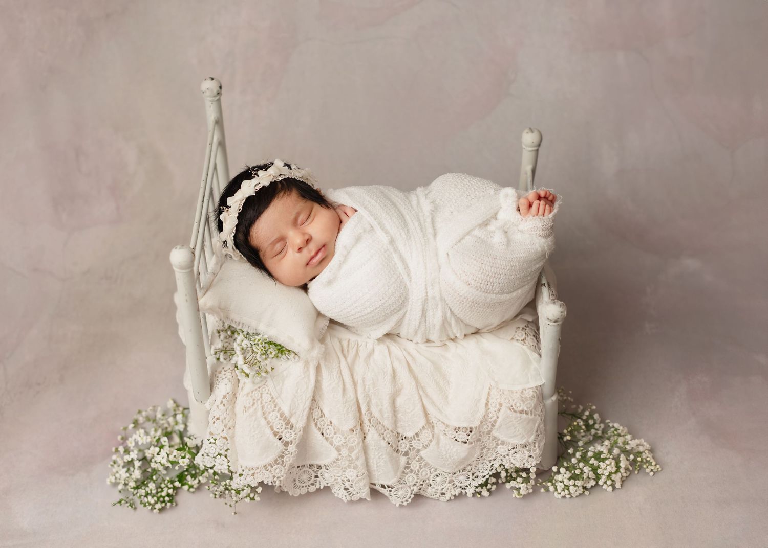 A newborn baby wearing white lace clothing lies sleeping peacefully on a soft gray backdrop with delicate baby's breath flowers. Elizabeth L