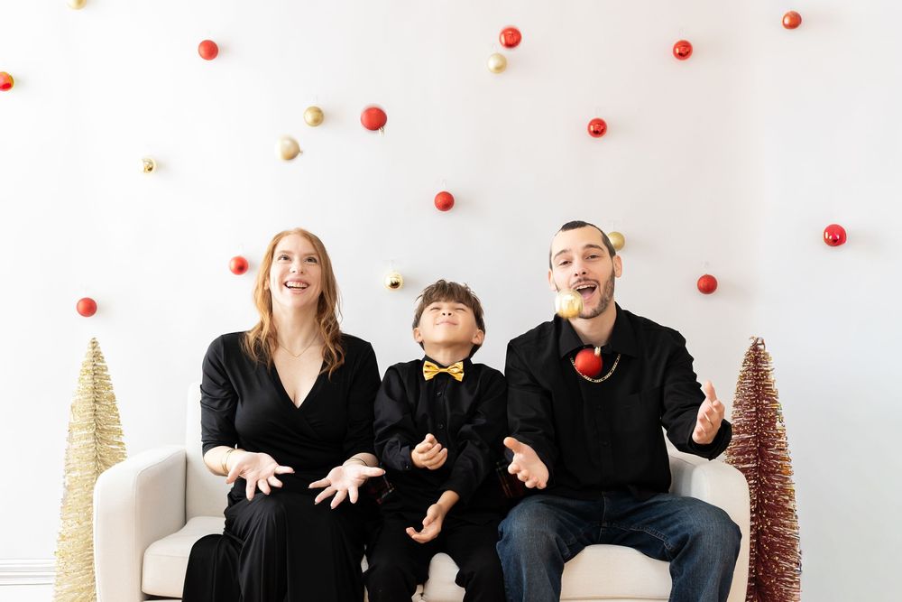 A group of people in black clothing toss red ornaments while sitting against a white background with holiday decorations.