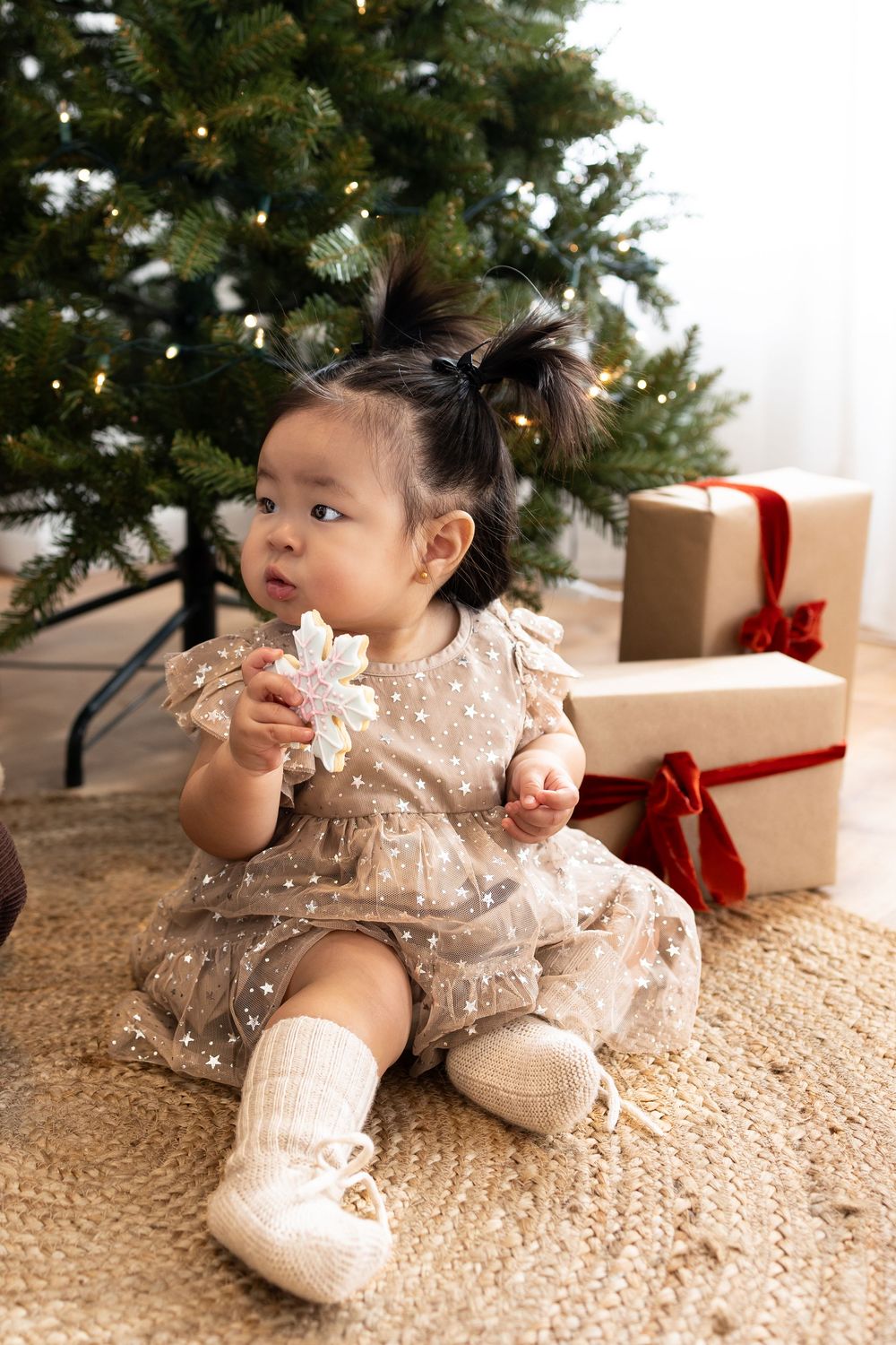 Baby in sparkly holiday dress sits on cream rug near Christmas tree with wrapped presents in background.