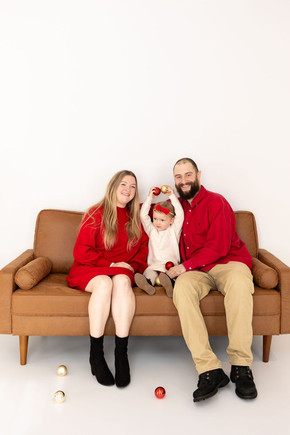Family in red and neutral clothing poses together on brown leather couch with Christmas ornaments.