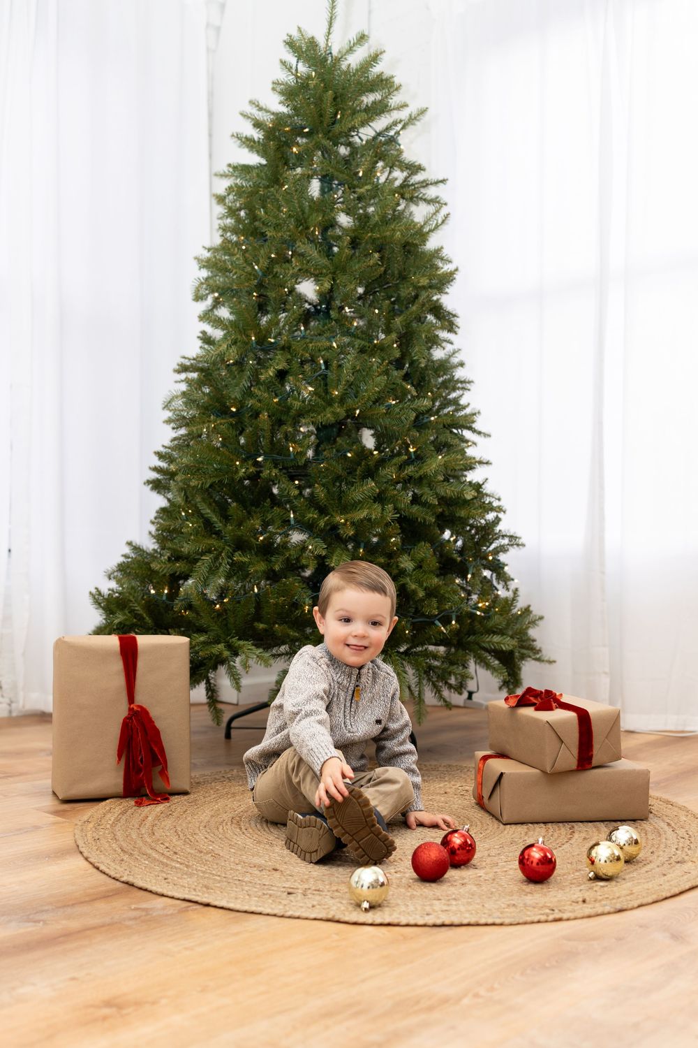 A young child sits on a jute rug by a decorated Christmas tree with red ornaments and wrapped presents.