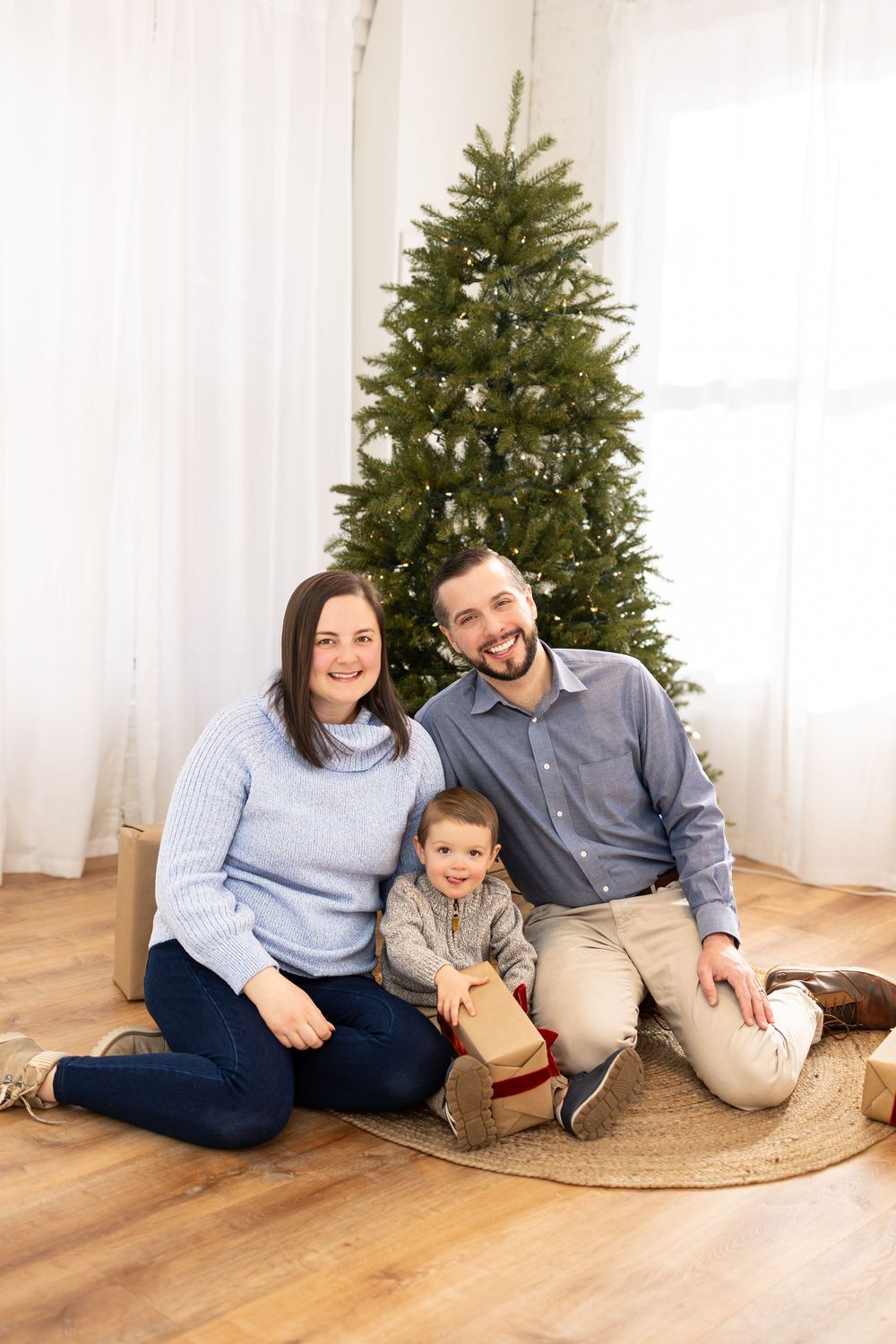Family relaxing together on wooden floor beside decorated Christmas tree in bright white room.