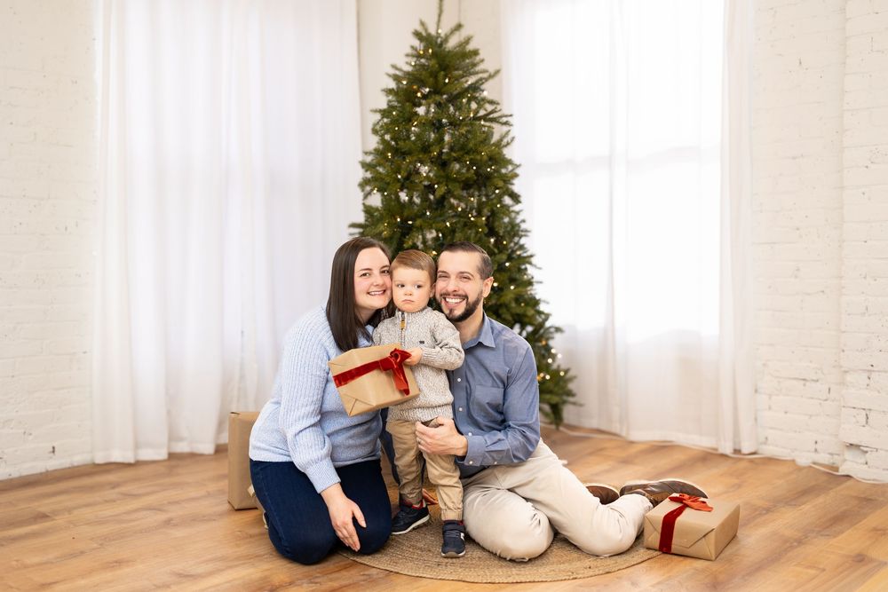 Family sitting on hardwood floor with decorated Christmas tree and white curtains in background.