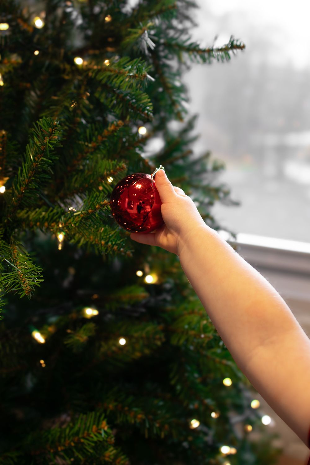 A hand reaches up to place a shiny red ornament on an illuminated Christmas tree with twinkling lights.