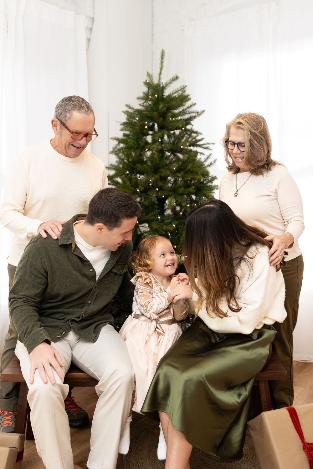 A family poses together in white and green outfits next to a Christmas tree for a holiday photo series.