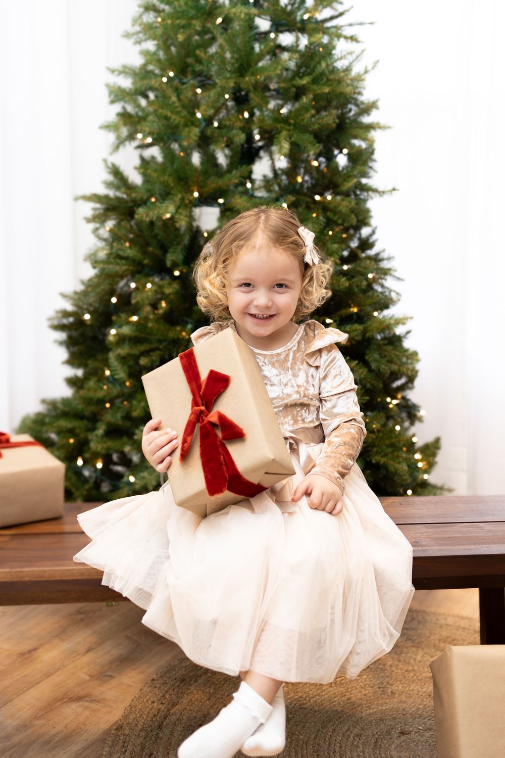 Child in white dress plays with Christmas presents on wooden bench near decorated tree.