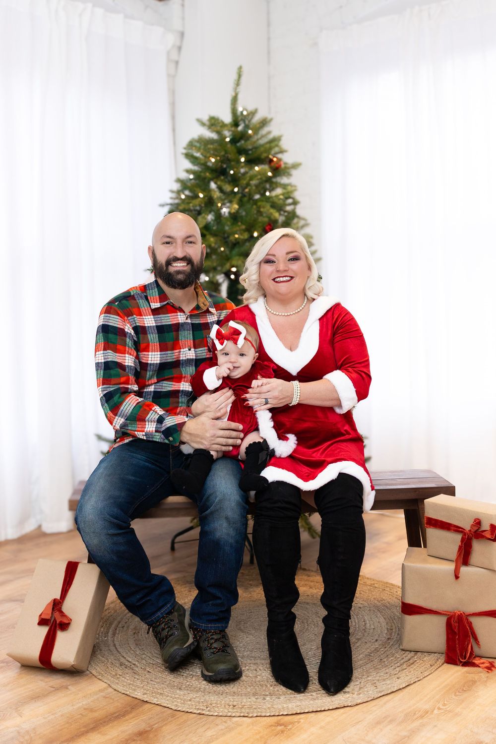 A couple in festive holiday attire laughs while sitting by a decorated Christmas tree with wrapped presents.