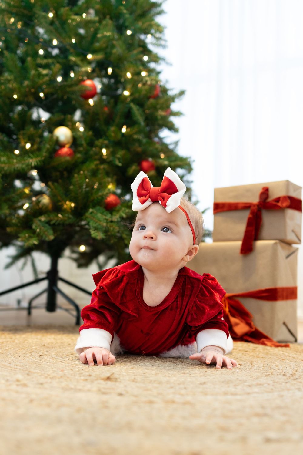 A baby in a red velvet dress crawls under a decorated Christmas tree near wrapped presents.