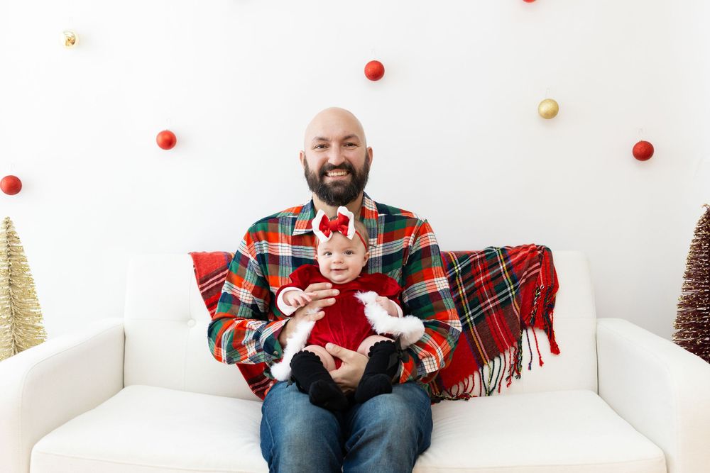 A person in a red plaid blanket sits on a white couch surrounded by festive holiday decor and small trees.