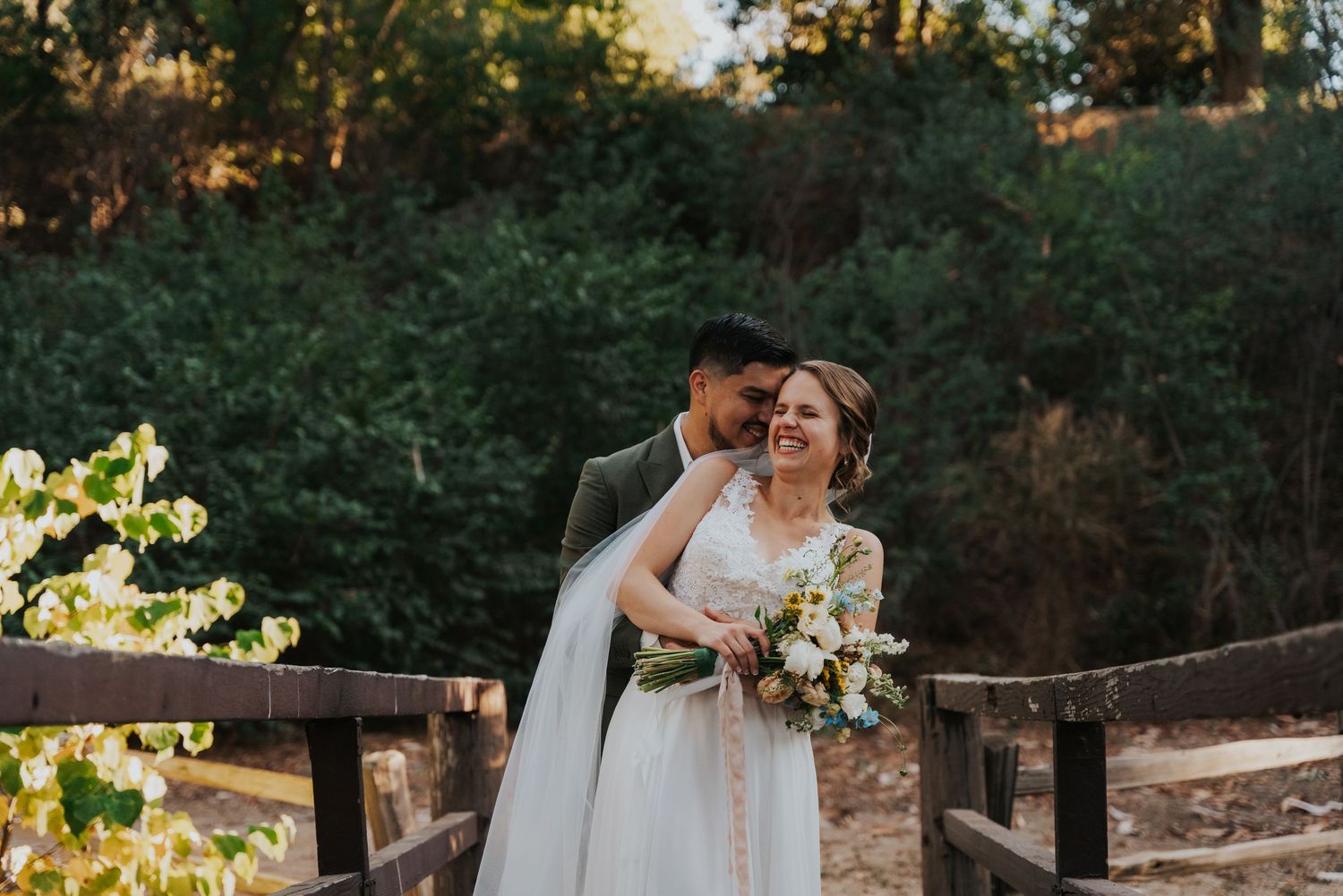 A romantic moment between newlyweds on a rustic wooden bridge during golden hour with white floral bouquet.