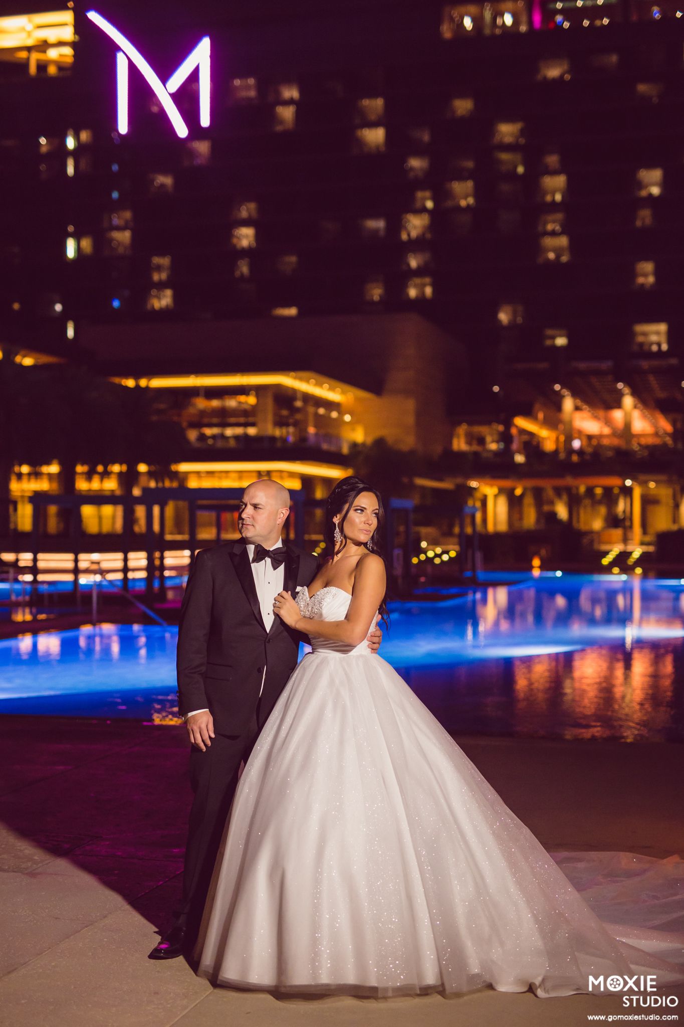 Bride in white ballgown and groom in black tuxedo pose romantically at night by illuminated pool with city skyline at M Resort.