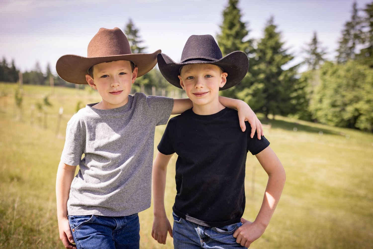 Two boys wearing cowboy hats pose together with arms around each other in a green field.