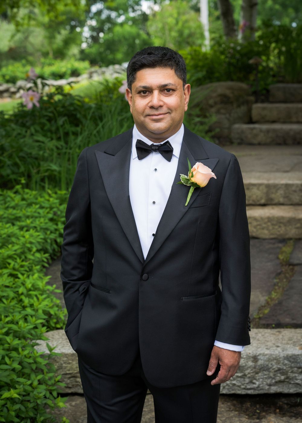 Groom in black tuxedo with bow tie and peach boutonniere stands before lush garden steps.