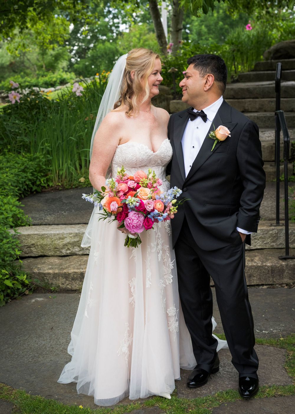 Bride in white lace gown holding colorful bouquet stands with groom in black tuxedo in garden wedding portrait.