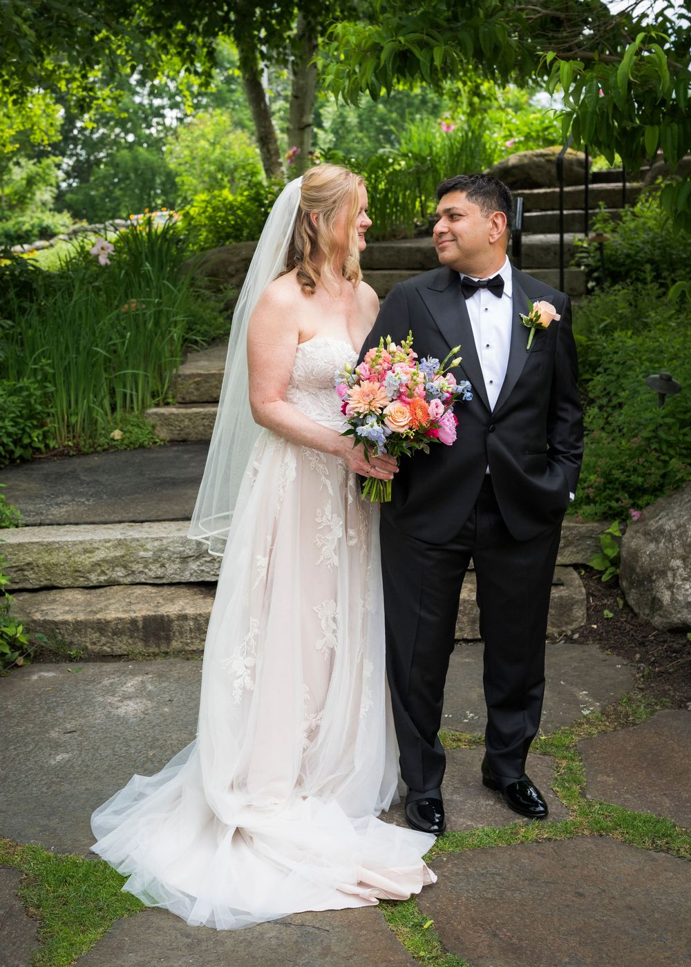 Bride and groom portrait in garden, she holds colorful bouquet wearing veil and white gown.