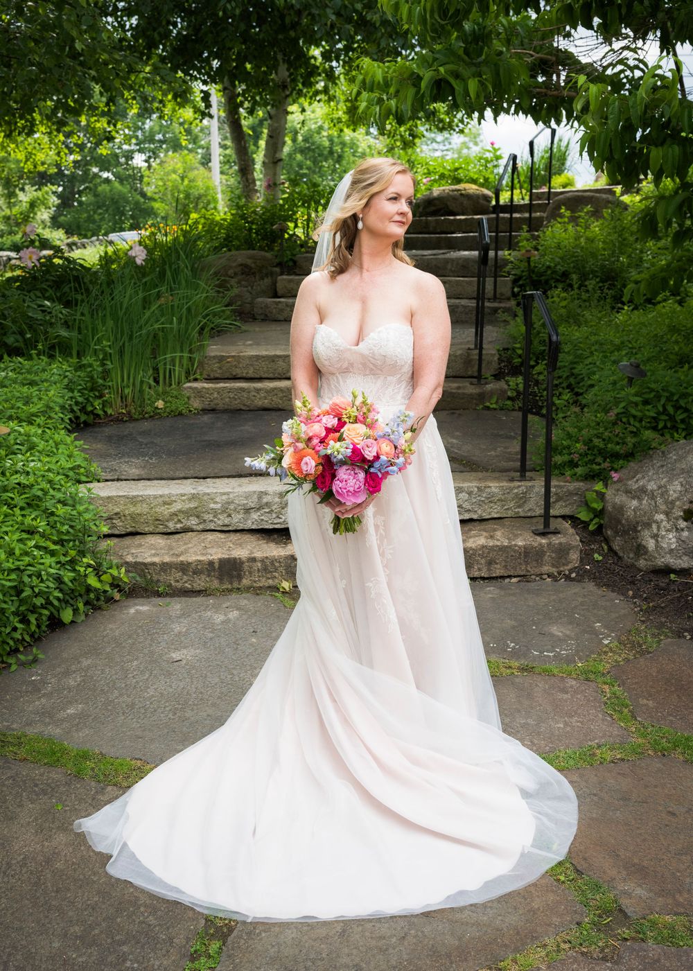 Elegant bride in a blush wedding gown holding a vibrant pink and orange bouquet in a lush garden setting.