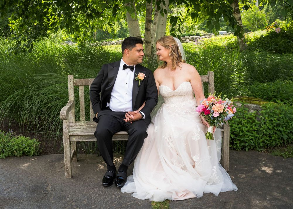 Bride and groom sitting on wooden bench in lush garden, bride holding colorful bouquet at wedding.