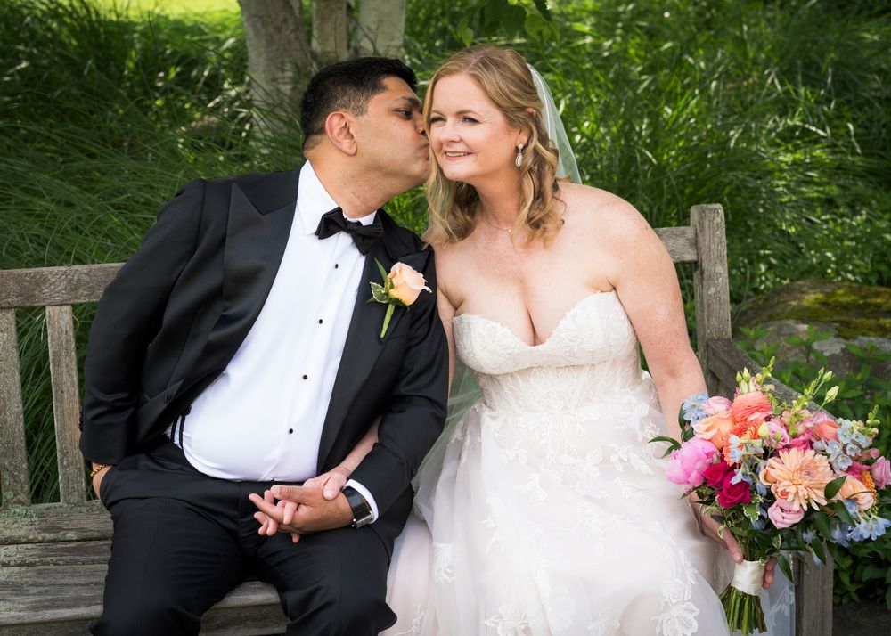 Groom kisses bride on cheek as they sit on bench, bride holding colorful floral bouquet.