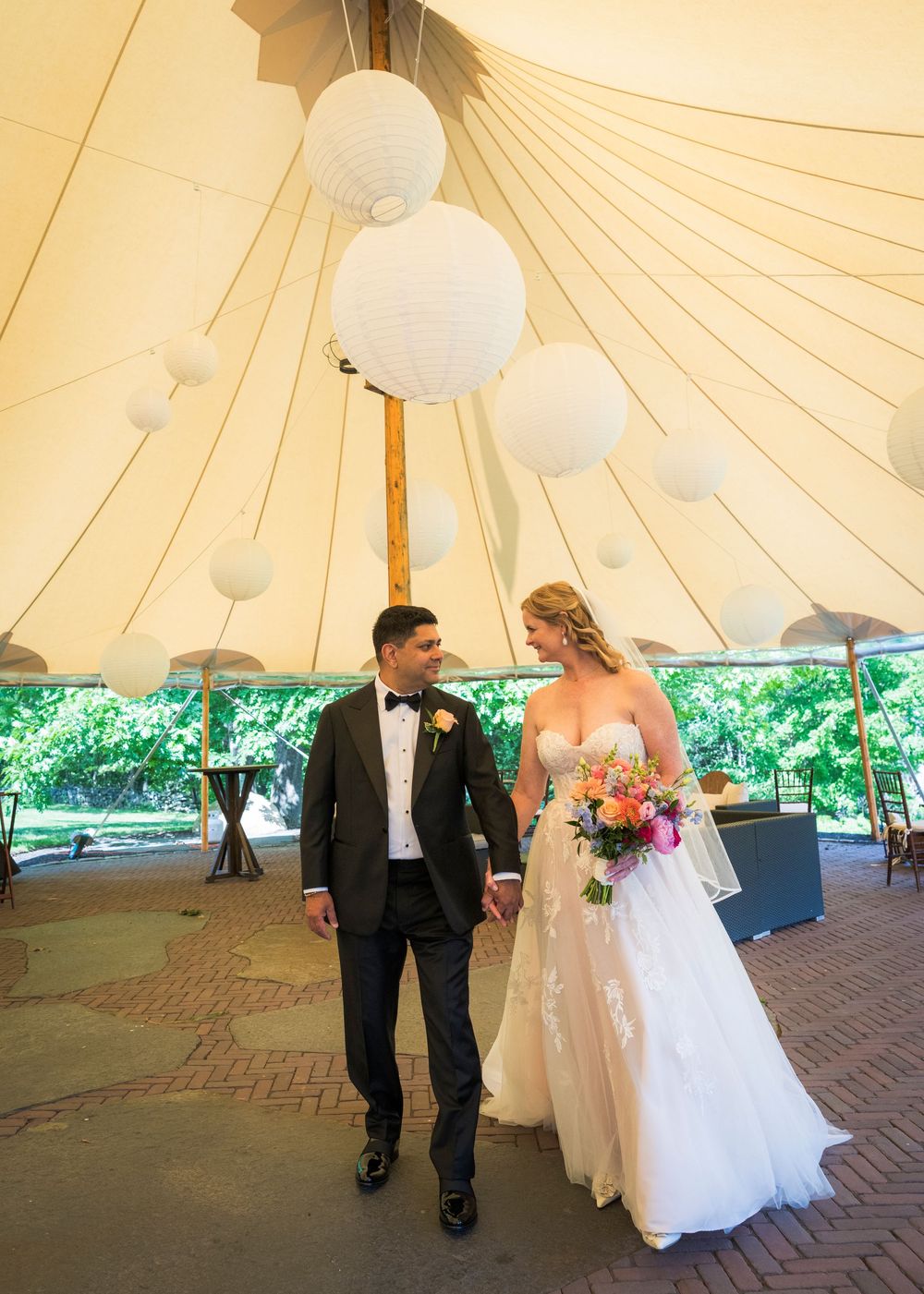 Bride and groom walk hand in hand under white paper lanterns inside elegant wedding tent.