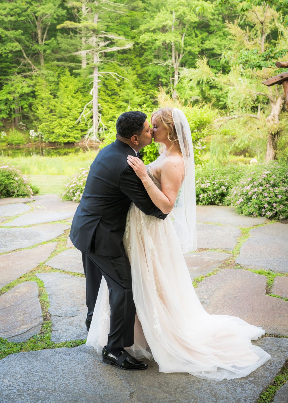 Bride and groom share romantic kiss on stone patio surrounded by lush green trees at outdoor wedding.