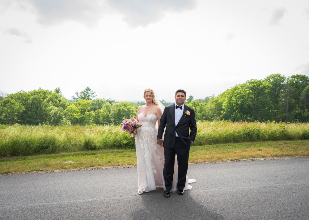 Bride in white gown and groom in tuxedo pose together on a path surrounded by lush greenery.