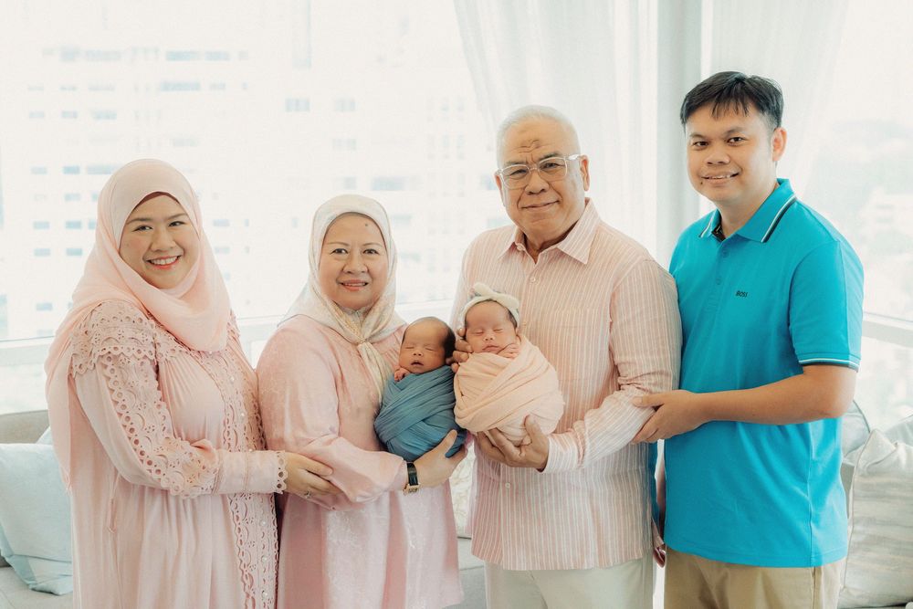 A multi-generational family in pastel colored clothing poses together while holding a baby in a bright indoor setting.