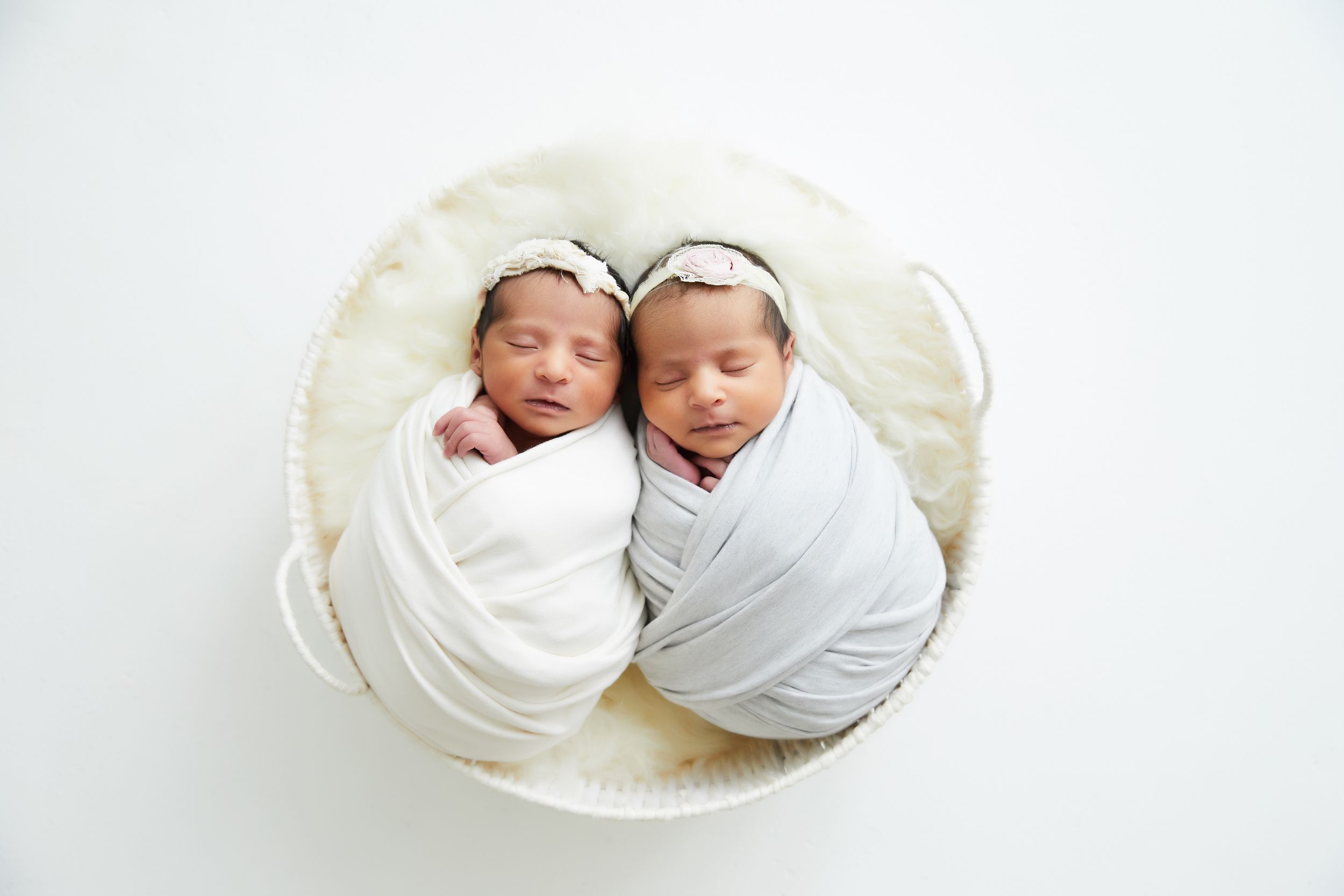 Two swaddled newborn babies sleeping together in a white round basket.