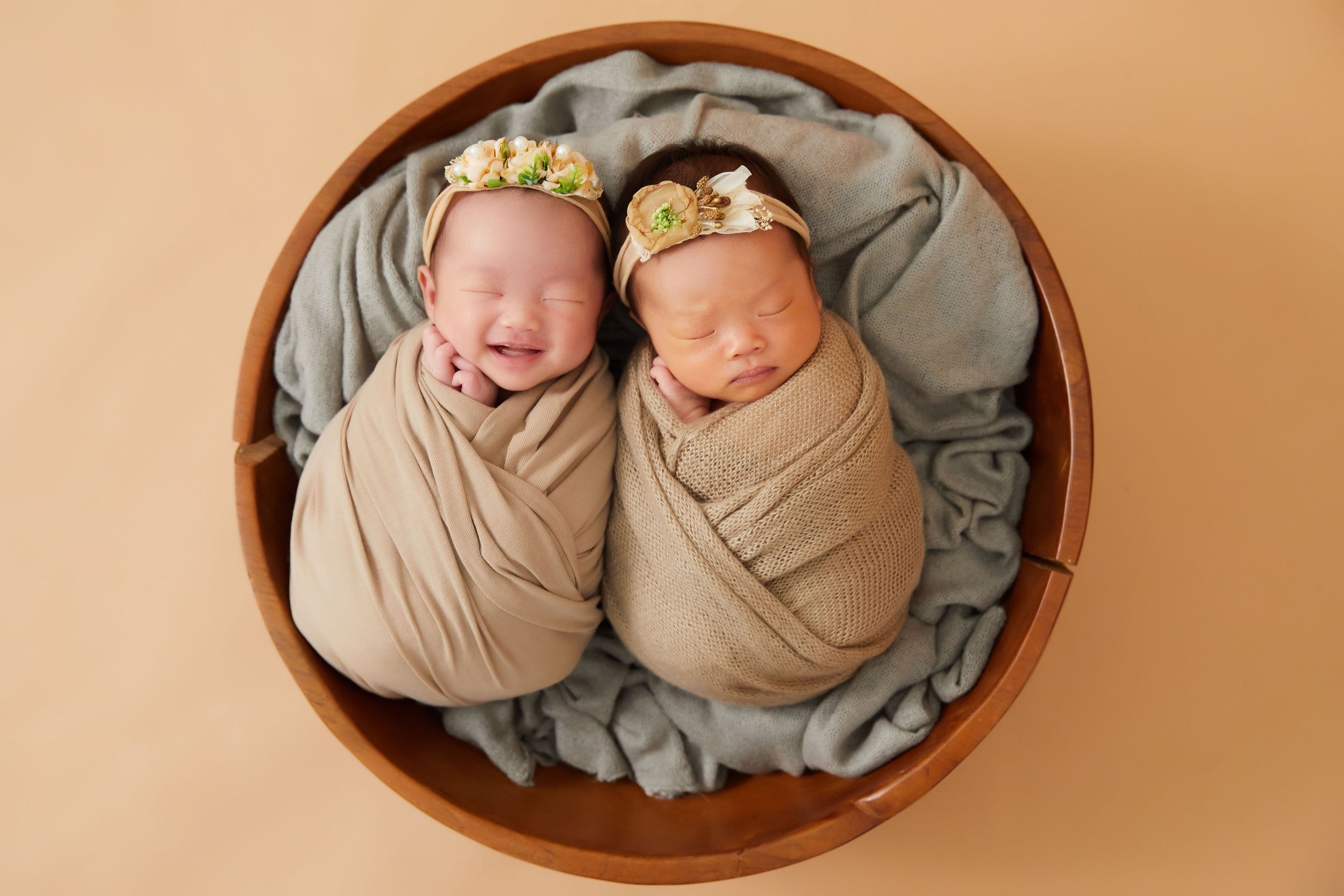 Two sleeping newborn babies swaddled in beige wraps wearing floral headbands lying in a round wooden bowl on gray fabric.
