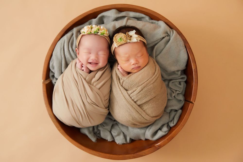 Two sleeping newborn babies swaddled in beige wraps wearing floral headbands lying in a round wooden bowl on gray fabric.