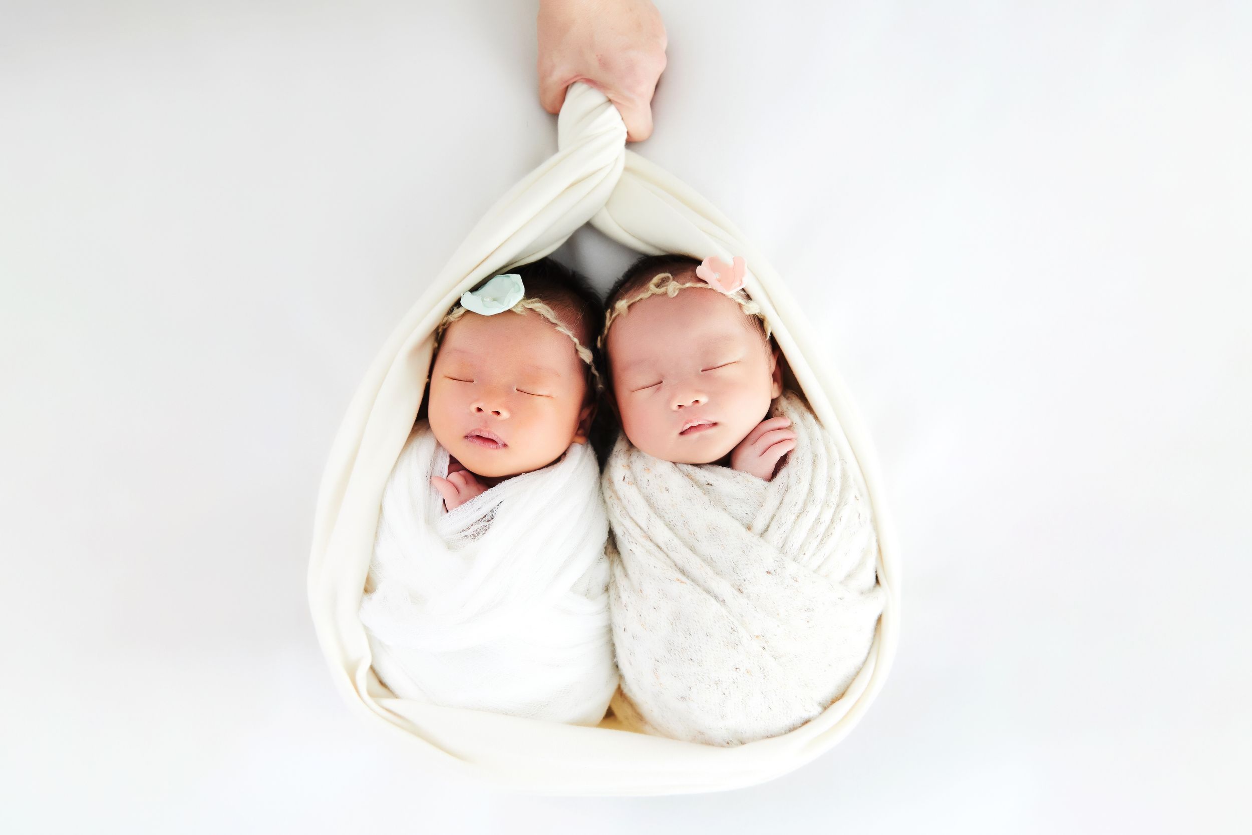 Two sleeping newborn babies swaddled in white blankets lying side by side on white background.