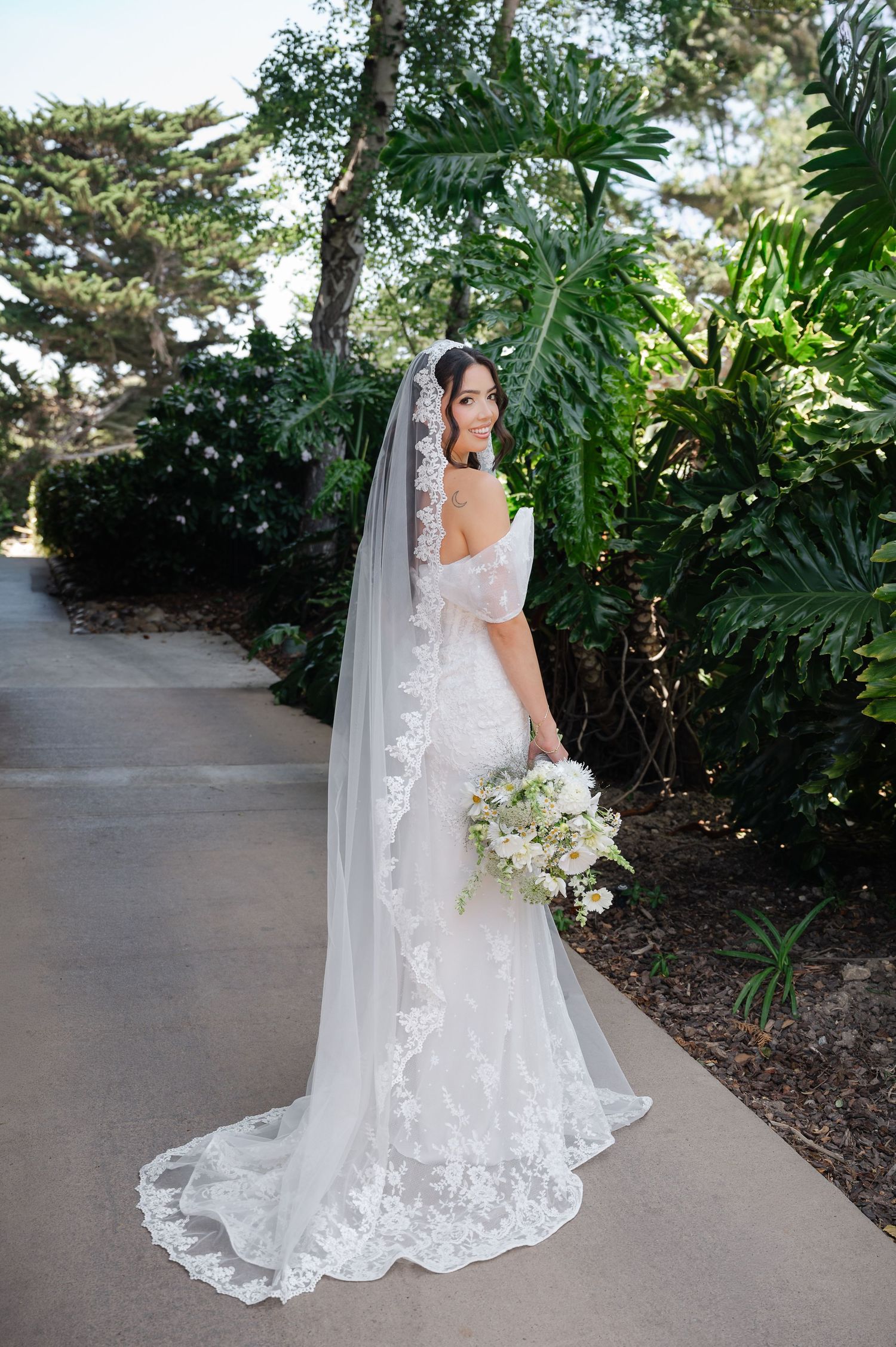 A bride in a white lace wedding gown and long veil holds an elegant white and green floral bouquet in a garden setting.