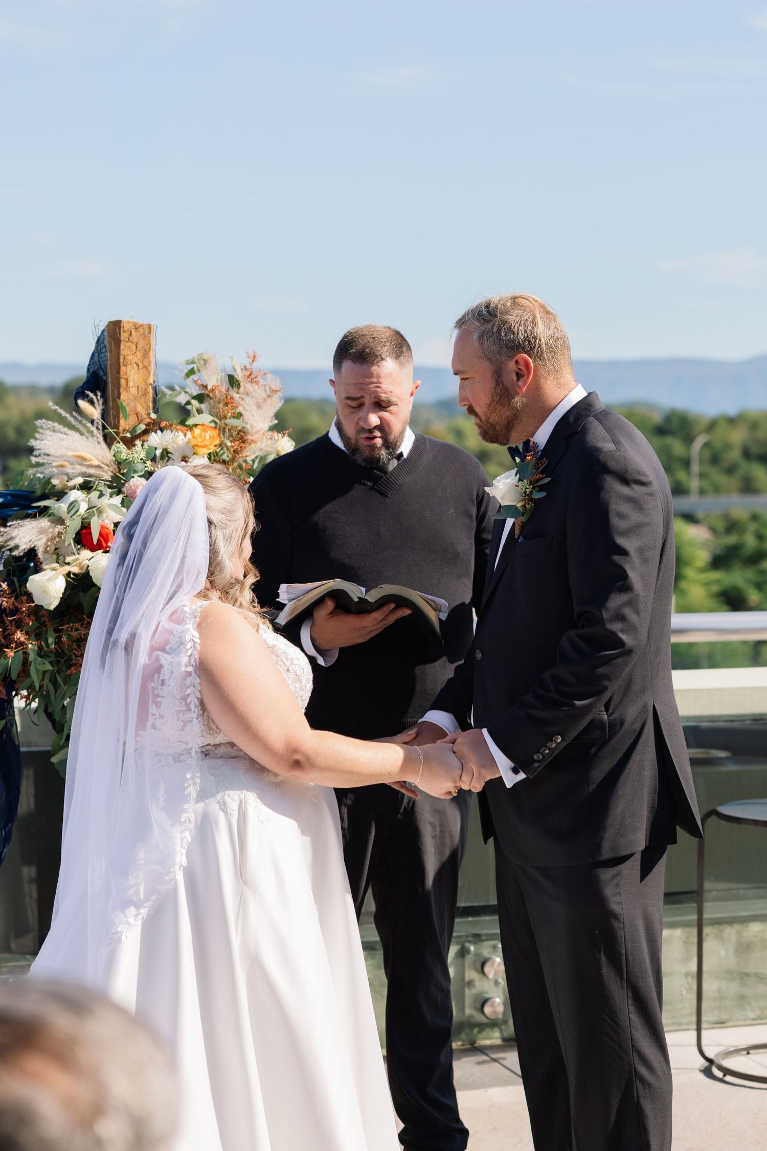 An outdoor wedding ceremony takes place on a balcony with a floral arch and scenic mountain views in the background.