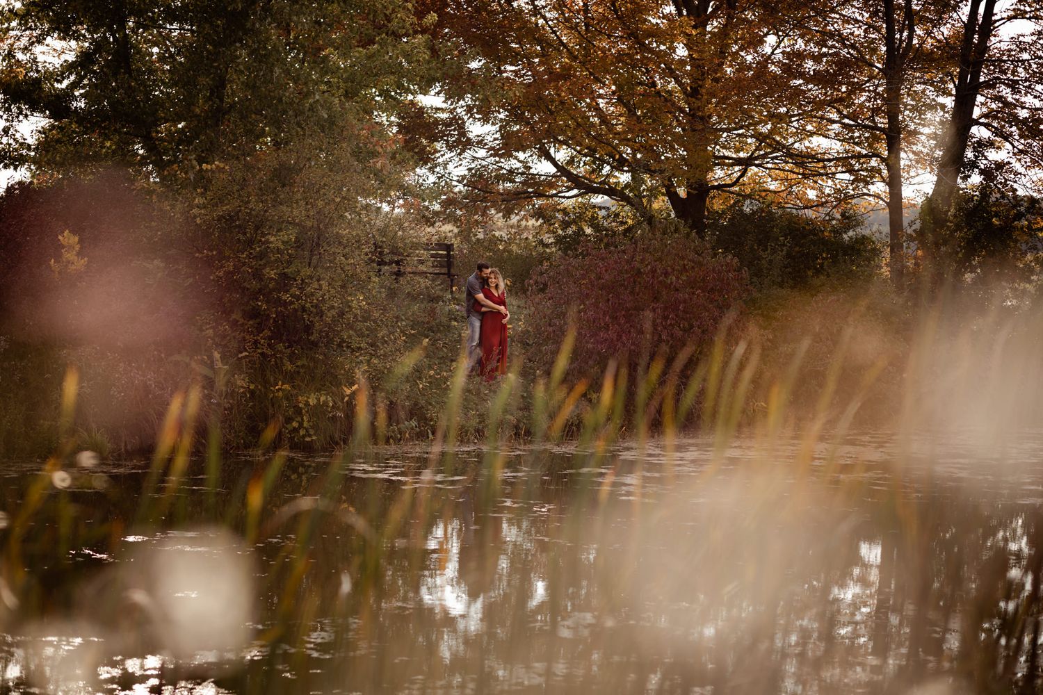 maternity couple hold baby belly while standing among fall foliage at cataraqui creek conservation area, kingston ontario