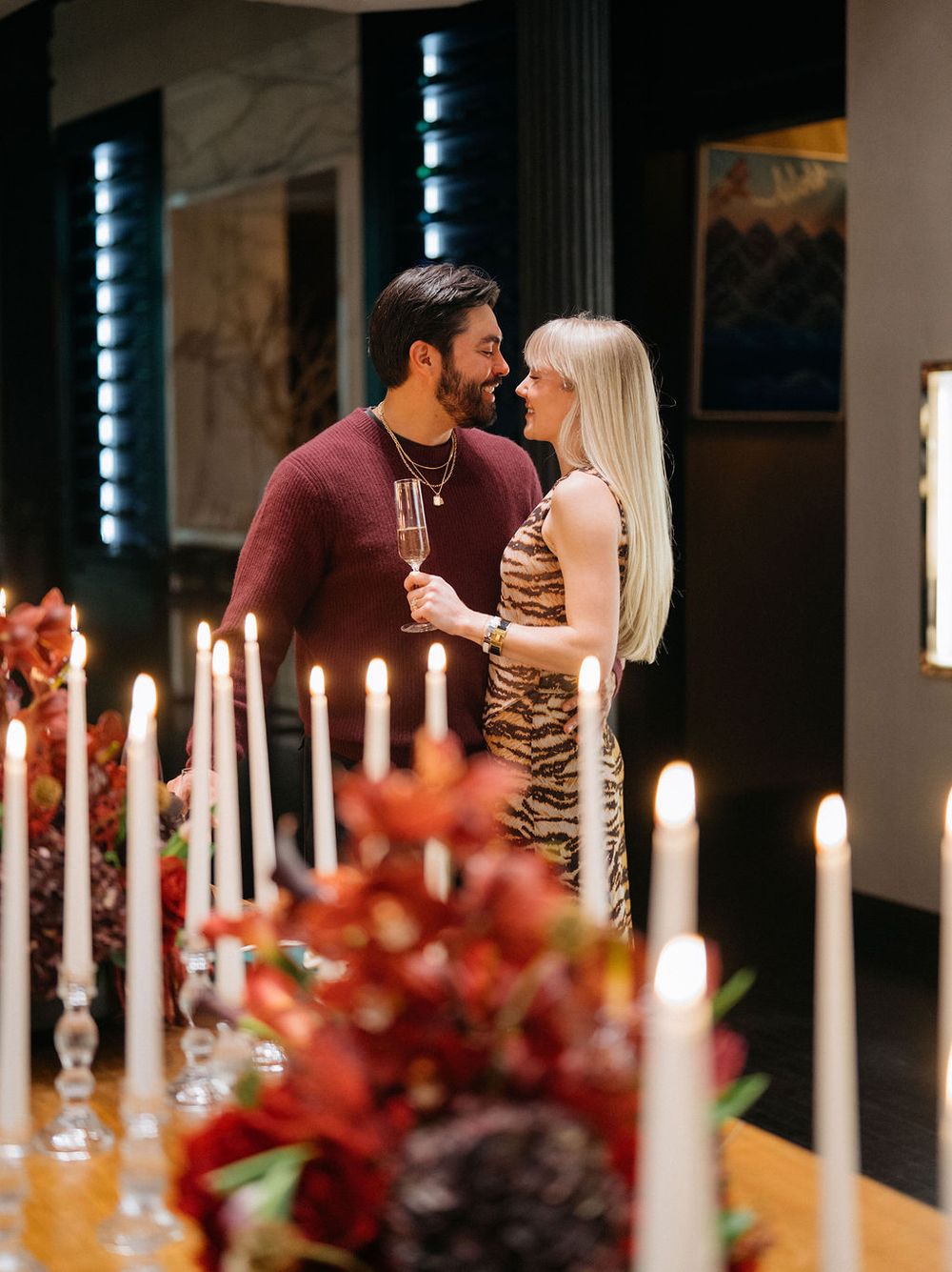 A romantic candlelit dinner table with red floral arrangements and white taper candles creates an intimate ambiance.