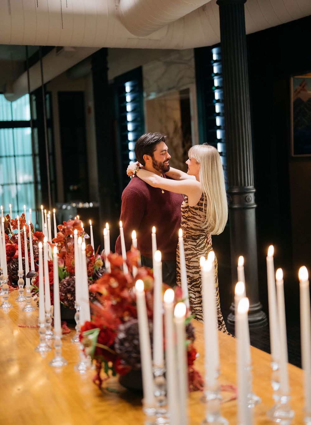 A romantic candlelit dinner table with white candles and red floral arrangements creates an intimate atmosphere.