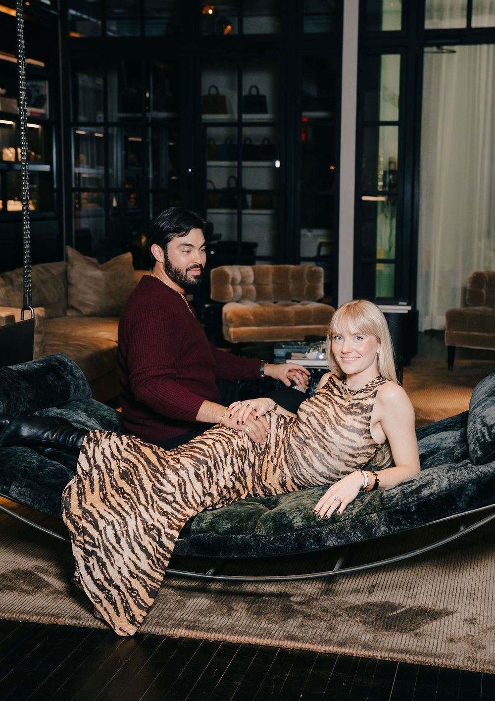 A couple lounges on a dark velvet chaise in an elegant room with dark wood and warm lighting.