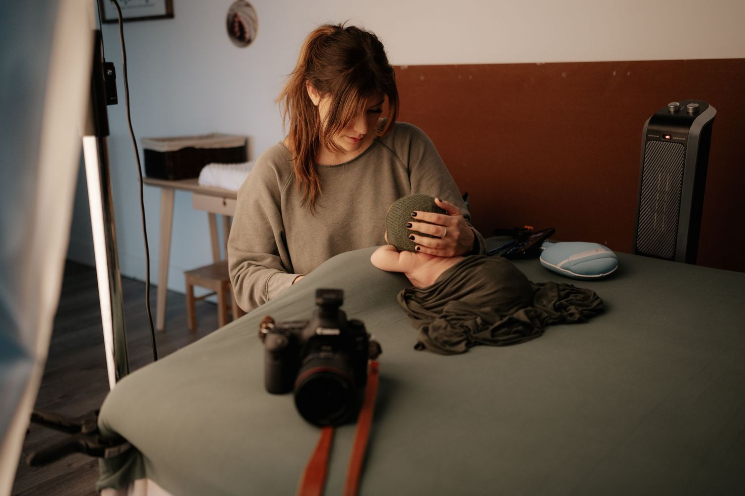 Someone in a beige sweater sits at a table with a camera and phone in a dimly lit room.